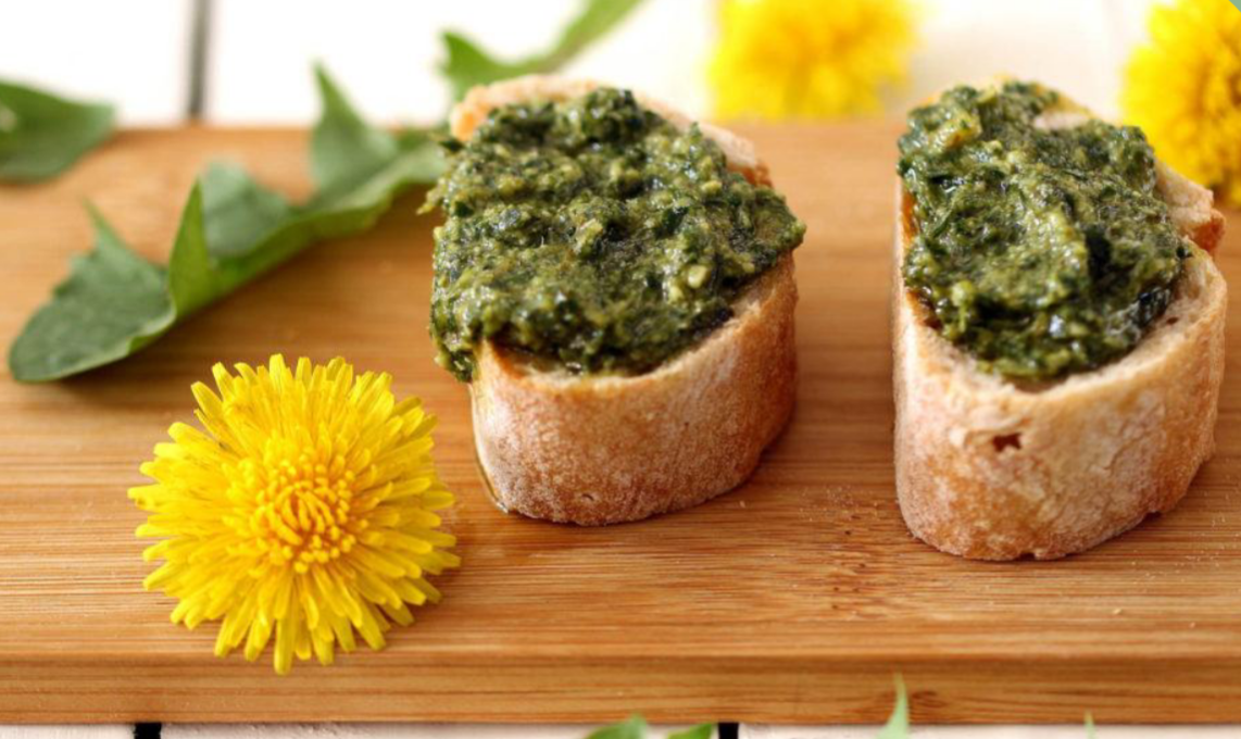 Bild von zwei mit Pesto beschmierten Scheiben eines Baguettes. Serviert auf einem Holzschneidebrett mit Löwenzahn als Dekoration.